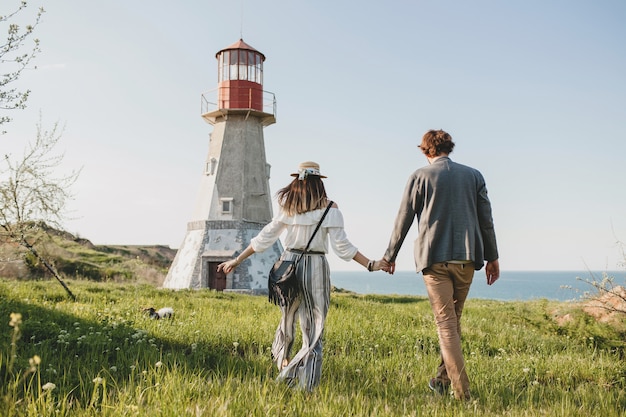 Vue de dos sur le style indie jeune couple hipster amoureux marcher dans la campagne, main dans la main, phare sur fond, chaude journée d'été, ensoleillée, tenue bohème