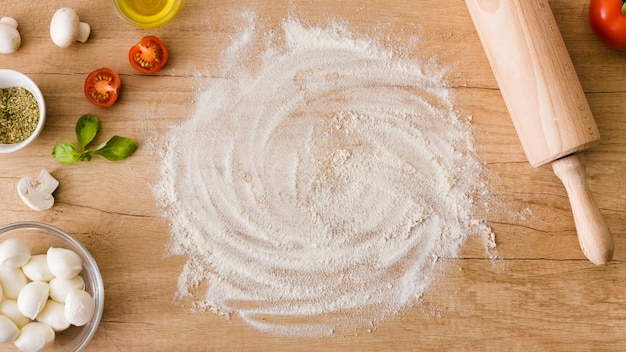 Photo gratuite une vue élevée de la farine avec des tomates; champignon; fromage basilic et mozzarella sur table en bois avec rouleau à pâtisserie sur table