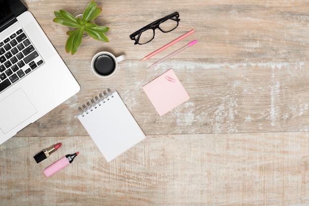 Vue élevée de l&#39;ordinateur portable; rouge à lèvres; surligneur; bloc-notes en spirale; tasse à café; stylo; et lunettes sur table en bois