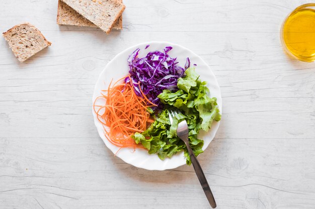 Vue élevée de salade saine avec une fourchette; tranches d&#39;huile et de pain contre un bureau en bois