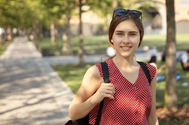 Vue Extérieure De La Belle Fille étudiante Mignonne Portant Haut En Pointillé Et Nuances Sur Sa Tête Portant Un Sac à Dos, Marchant Dans Le Parc Après L'université. Femme élégante Ayant Marcher. Concept De Personnes Et De Style De Vie