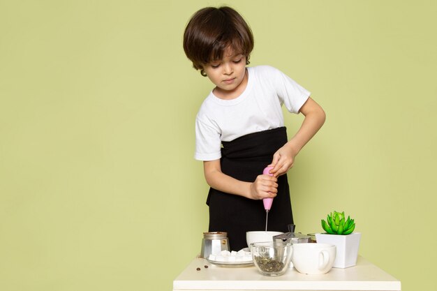 Une vue de face adorable garçon mignon en t-shirt blanc prépare du café sur la table sur l'espace de couleur pierre