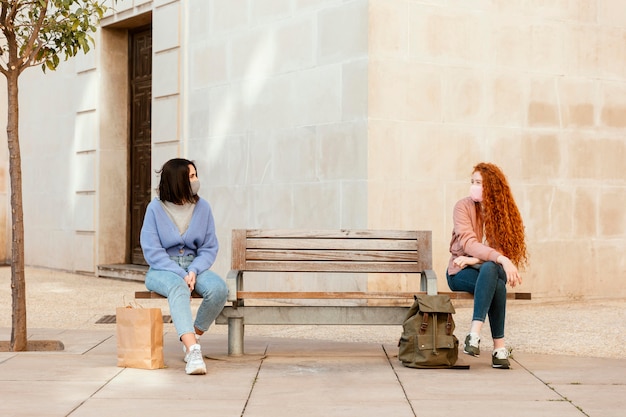 Vue de face des amies avec des masques faciaux à l'extérieur assis sur un banc