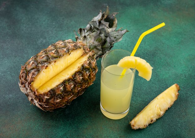 Vue de face de l'ananas avec un morceau coupé de fruits entiers et de jus d'ananas sur une surface verte