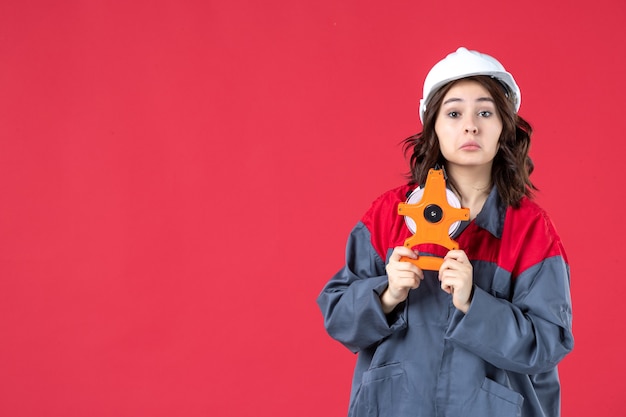 Vue de face de l'architecte curieux en uniforme avec casque tenant un ruban à mesurer sur un mur rouge isolé