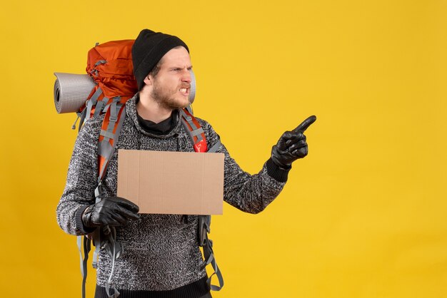 Vue de face de l'auto-stoppeur masculin en colère avec des gants en cuir et sac à dos tenant un carton vierge pointant vers la bonne direction