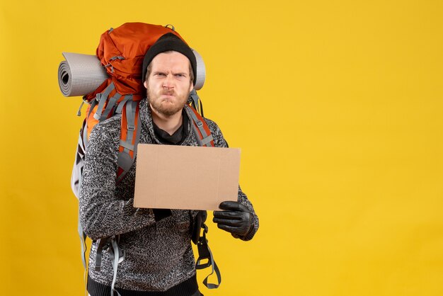 Vue de face de l'auto-stoppeur masculin insatisfait avec des gants en cuir et sac à dos tenant un carton vierge