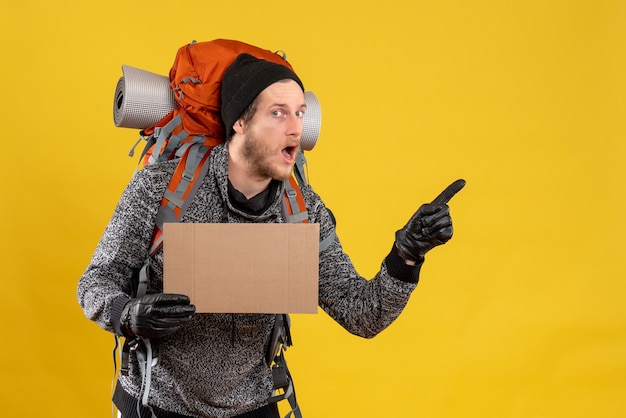 Vue de face de l'auto-stoppeur masculin intéressé avec des gants en cuir et sac à dos tenant un carton vierge pointant vers quelque chose