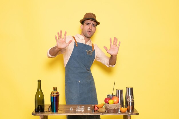 Vue de face barman masculin devant la table avec des boissons et des shakers sur le mur jaune nuit alcool bar club boisson masculine