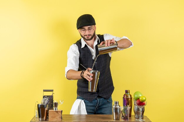 Vue de face barman masculin devant la table avec des shakers faisant un verre sur un bar mural jaune alcool nuit club de boissons pour les jeunes