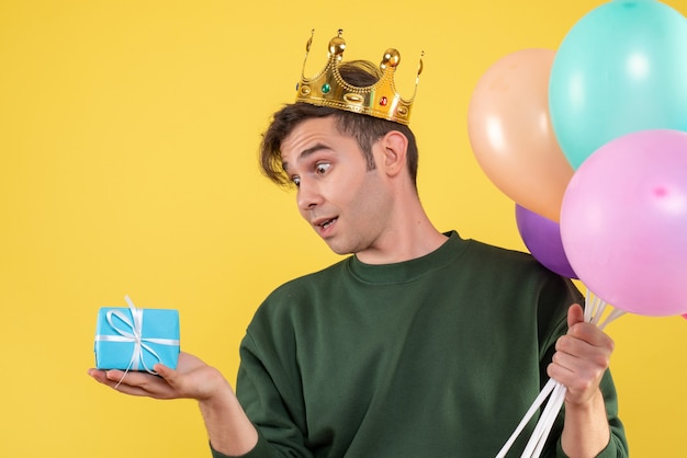 Vue de face beau jeune homme avec couronne tenant des ballons regardant cadeau sur jaune