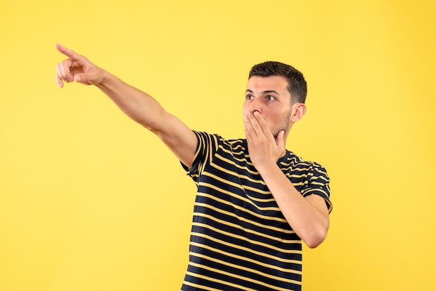 Vue de face beau mâle en t-shirt rayé noir et blanc pointant avec le doigt quelque chose sur fond isolé jaune