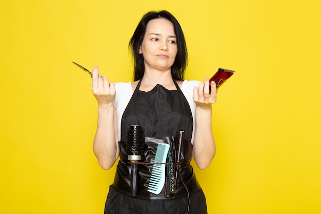 Une vue de face belle femme coiffeuse en t-shirt blanc cape noire avec des brosses avec des cheveux lavés tenant des ciseaux et une machine posant sur le fond jaune coiffeur coiffeur