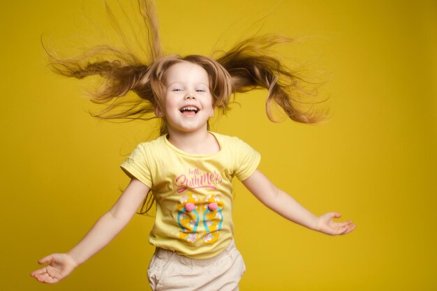 Vue de face de la belle fille aux cheveux longs en chemise mignonne jouant avec les cheveux et virevoltant en studio Petit enfant ensoleillé regardant la caméra et posant sur fond isolé jaune Concept de l'enfance