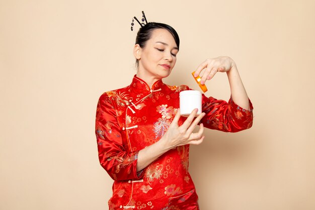 Une vue de face belle geisha japonaise en robe japonaise rouge traditionnelle avec des bâtons de cheveux posant la crème d'ouverture peut sourire sur la cérémonie de fond crème Japon