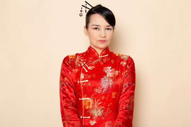 Une vue de face belle geisha japonaise en robe japonaise rouge traditionnelle avec des bâtons de cheveux posant debout sur la cérémonie de fond crème divertissant le Japon