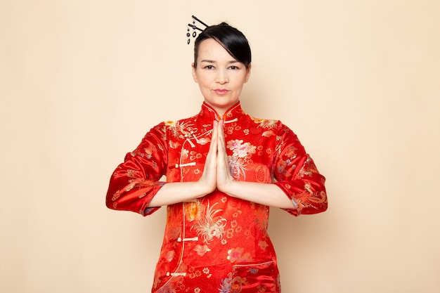 Photo gratuite une vue de face belle geisha japonaise en robe japonaise rouge traditionnelle avec des bâtons de cheveux posant avec ses mains debout sur la cérémonie de fond crème divertissant le japon est