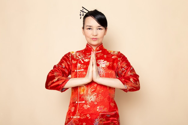 Une vue de face belle geisha japonaise en robe japonaise rouge traditionnelle avec des bâtons de cheveux posant avec ses mains debout sur la cérémonie de fond crème divertissant le Japon est