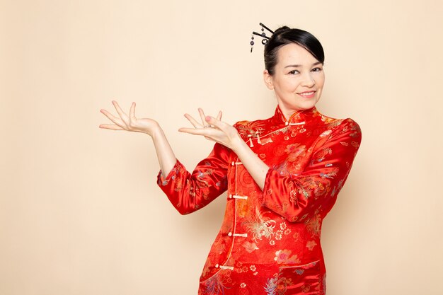 Une vue de face belle geisha japonaise en robe japonaise rouge traditionnelle avec des bâtons de cheveux posant avec ses mains debout souriant sur la cérémonie de fond crème divertissant le Japon est