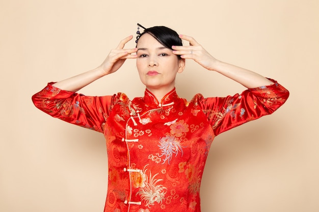 Une vue de face belle geisha japonaise en robe japonaise rouge traditionnelle avec des bâtons de cheveux posant avec ses mains élégantes sur la cérémonie de fond crème divertissant le Japon est