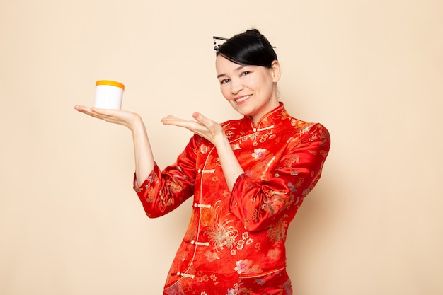 Une vue de face belle geisha japonaise en robe japonaise rouge traditionnelle avec des bâtons de cheveux posant tenant la crème peut sourire sur la cérémonie de fond crème Japon