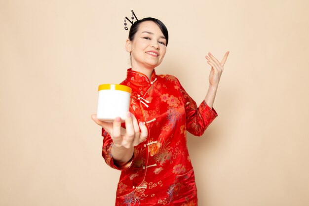 Une vue de face belle geisha japonaise en robe japonaise rouge traditionnelle avec des bâtons de cheveux posant tenant la crème peut sourire sur la cérémonie de fond crème Japon