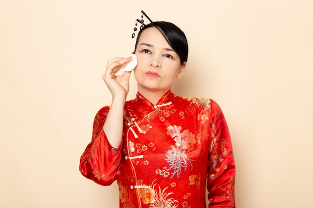 Photo gratuite une vue de face belle geisha japonaise en robe japonaise rouge traditionnelle avec des bâtons de cheveux posant tenant peu de coton blanc nettoyer son visage sur la cérémonie de fond crème japon