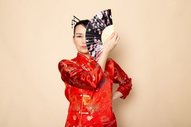 Une vue de face belle geisha japonaise en robe japonaise rouge traditionnelle avec des bâtons de cheveux posant tenant un ventilateur pliant élégant sur la cérémonie de fond crème Japon