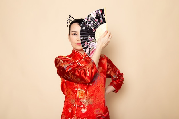 Une vue de face belle geisha japonaise en robe japonaise rouge traditionnelle avec des bâtons de cheveux posant tenant un ventilateur pliant élégant sur la cérémonie de fond crème Japon