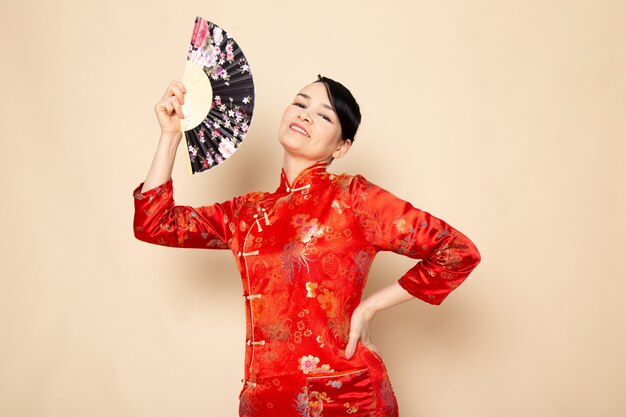 Une vue de face belle geisha japonaise en robe japonaise rouge traditionnelle avec des bâtons de cheveux posant tenant un ventilateur pliant souriant sur la cérémonie de fond crème Japon