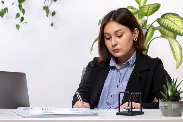 Une vue de face belle jeune femme d'affaires en veste noire et chemise bleue travaillant avec un ordinateur portable en écrivant les notes devant le bureau de l'emploi des entreprises de table