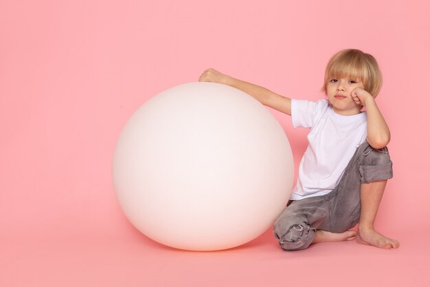 Une vue de face blond mignon garçon en t-shirt blanc jouant avec une boule blanche ronde sur l'espace rose