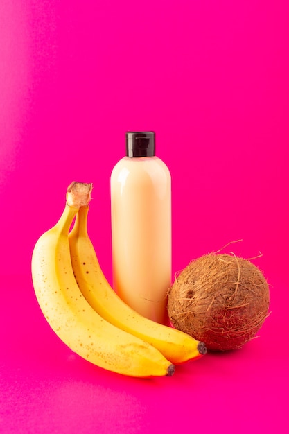 Une vue de face bouteille de shampooing en plastique de couleur crème peut avec capuchon noir isolé avec des bananes et de la noix de coco sur les cheveux de beauté cosmétiques fond rose