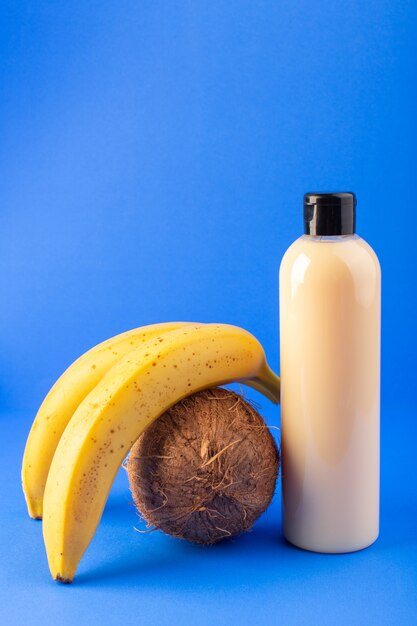 Une vue de face bouteille de shampooing en plastique de couleur crème peut avec capuchon noir isolé avec de la noix de coco et des bananes sur le fond bleu cheveux beauté cosmétiques