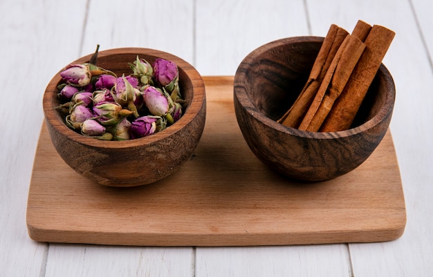 Vue de face de la cannelle avec des boutons de rose séchés dans des bols sur une planche sur une surface blanche