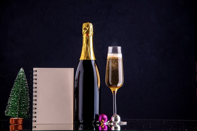 Vue de face champagne en bouteille et mini cahier d'arbre de Noël en verre sur une surface sombre