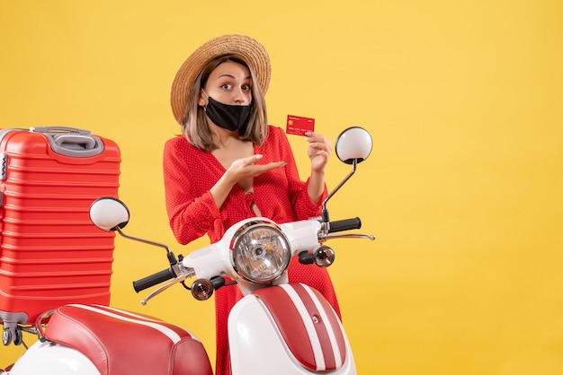 Vue de face de la charmante femme avec masque noir tenant une carte de crédit près de cyclomoteur