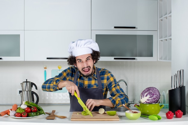 Vue de face d'un chef masculin effrayé avec des légumes frais et cuisine avec des ustensiles de cuisine et hacher des poivrons verts dans la cuisine blanche