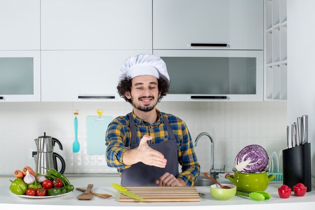 Vue de face d'un chef masculin émotionnel avec des légumes frais et cuisine avec des ustensiles de cuisine et accueillant quelqu'un dans la cuisine blanche