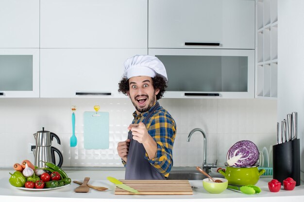 Vue de face d'un chef masculin heureux et positif avec des légumes frais et cuisine avec des ustensiles de cuisine et dans la cuisine blanche