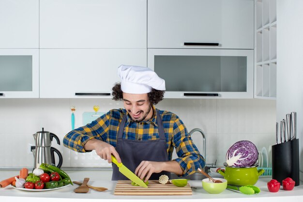 Vue de face d'un chef masculin positif avec des légumes frais et cuisine avec des ustensiles de cuisine et hacher des poivrons verts dans la cuisine blanche