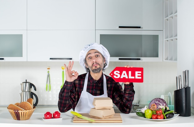Photo gratuite vue de face d'un chef masculin satisfait en uniforme brandissant un panneau de vente rouge faisant des gestes ok dans une cuisine moderne
