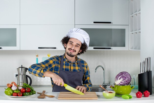 Vue de face d'un chef masculin souriant avec des légumes frais et cuisinant avec des ustensiles de cuisine et coupant quelque chose dans la cuisine blanche