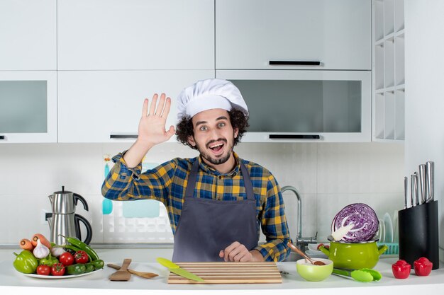 Vue De Face D'un Chef Masculin Souriant Avec Des Légumes Frais Et Cuisinant Avec Des Ustensiles De Cuisine Et Disant Bonjour Dans La Cuisine Blanche