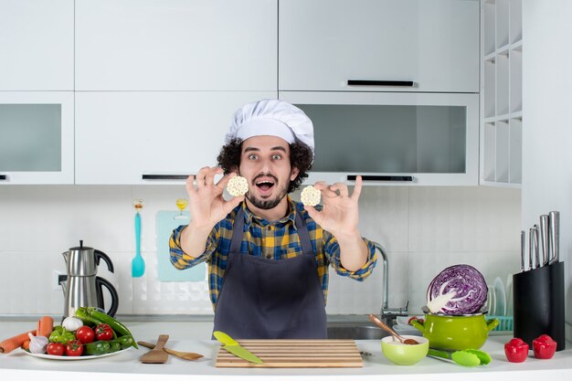 Vue de face d'un chef masculin souriant avec des légumes frais et cuisine avec des ustensiles de cuisine et montrant la nourriture dans la cuisine blanche