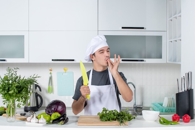 Vue de face chef masculin en uniforme tenant un couteau dans la cuisine faisant un baiser de chef