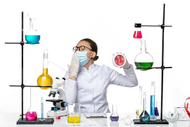 Vue de face chimiste femme en costume médical avec masque tenant des horloges bâillant sur le fond blanc virus laboratoire chimie covid- splash