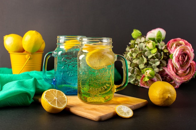 Une vue de face cocktail de citron boisson fraîche fraîche à l'intérieur de tasses en verre tranché et citrons entiers avec des fleurs sur le fond sombre boisson cocktail fruit