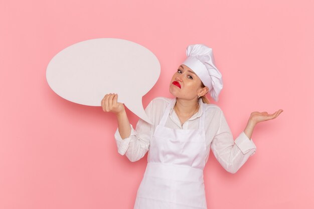 Vue de face confiseur femme en vêtements blancs posant avec panneau blanc sur le travail de pâtisserie sucrée confiserie mur rose