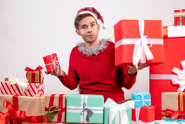 Vue de face confus jeune homme assis autour de cadeaux de Noël